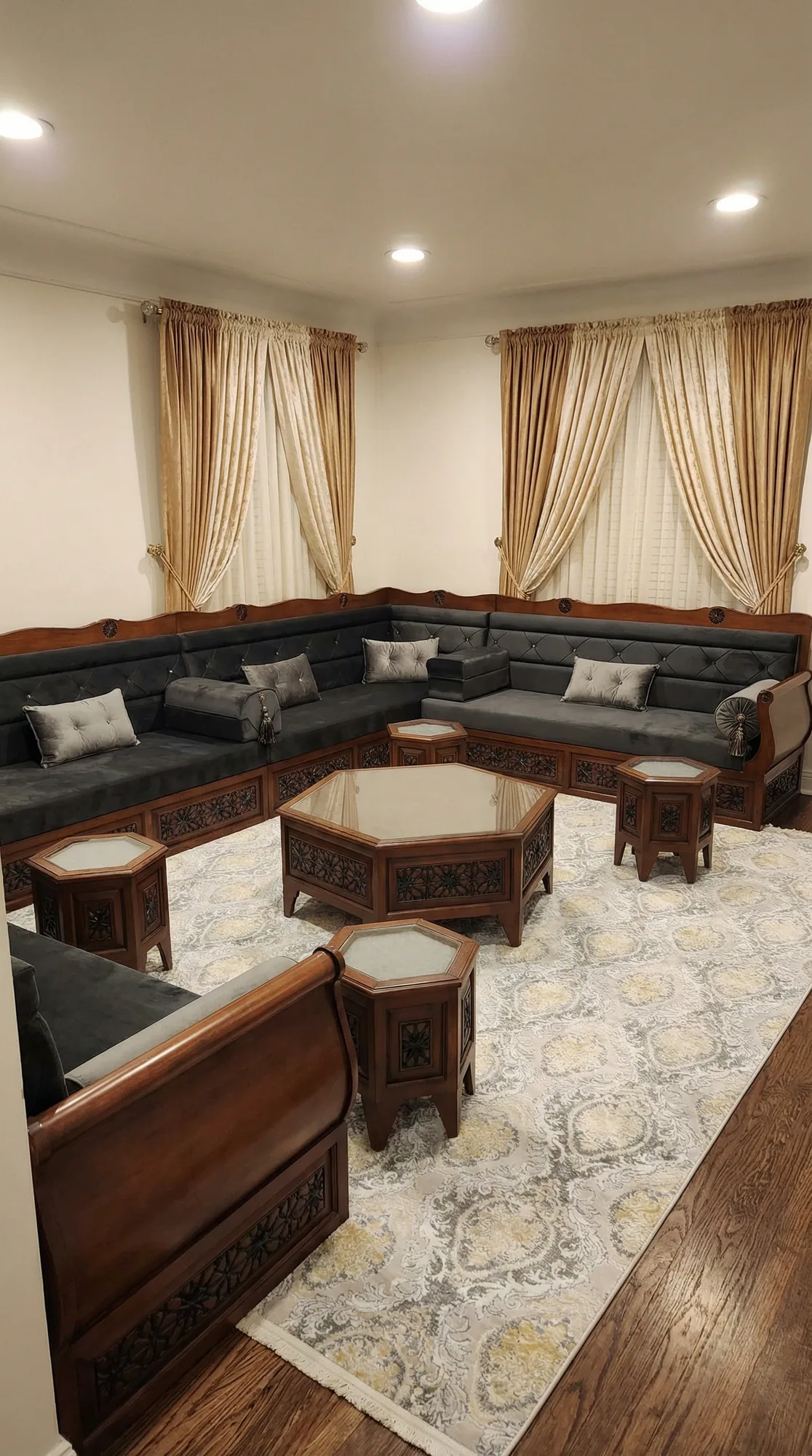 Carved walnut-framed Arabic majlis with tufted charcoal cushions along three walls and hexagonal wooden tables on a cream rug