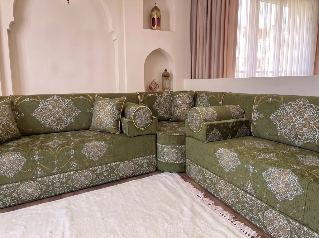 Low corner view of a khaki green medallion majlis sofa with gold-piped bolster armrests and a white fringed rug
