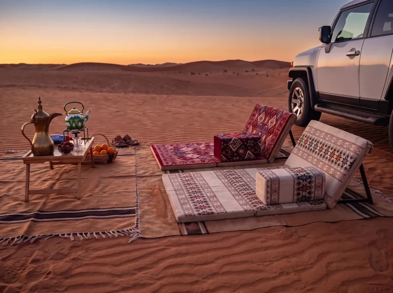 Two folding Arabic sunbed loungers, maroon and beige, on tan striped rugs at desert sunset beside a 4x4