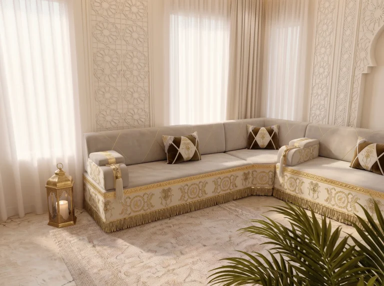 Corner of a beige Arabic majlis with gold-embroidered cream bases, framed by a brass lantern and palm fronds in front