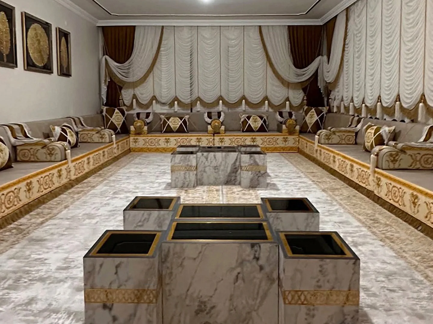 Beige Arabic majlis with gold baroque-embroidered bases along three walls, marble cube tables and white festoon curtains