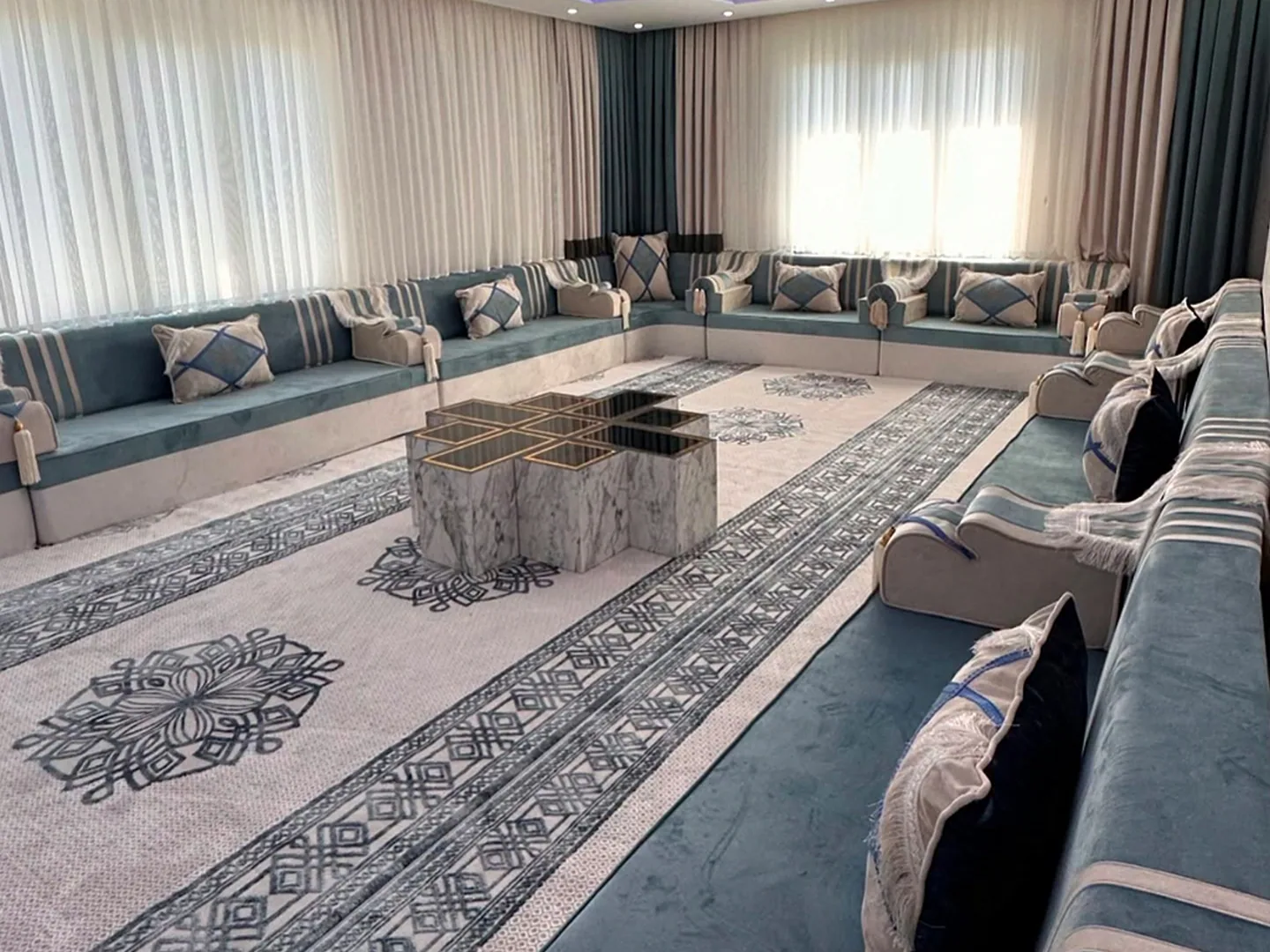 Dusty blue velvet Arabic majlis with cream-striped backs along three walls, a marble cross-shaped table and a blue-banded rug