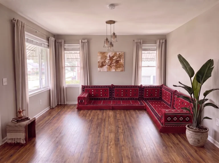 Red sadu-weave Arabic floor sofa in an L shape along two grey walls, with a bare wooden floor and a tall potted plant