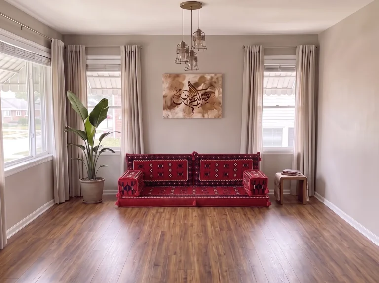 Red Arabic floor sofa in sadu weave centred between two windows under a calligraphy canvas, floor left bare