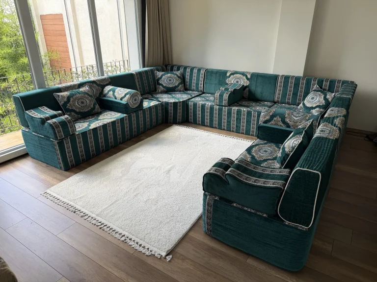 Teal velvet Arabic majlis in a U shape on a wooden floor beside a balcony window, white fringed rug