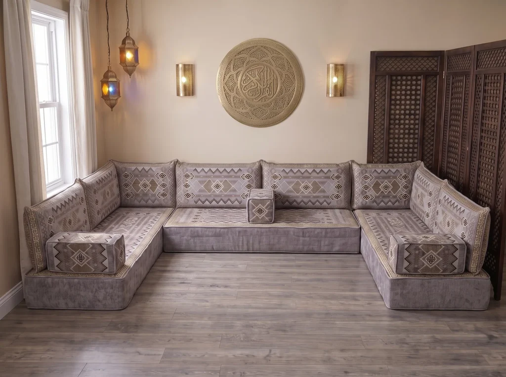 Thick gray and gold kilim-pattern Arabic floor couch in a U shape on a bare wood floor below a brass wall plate